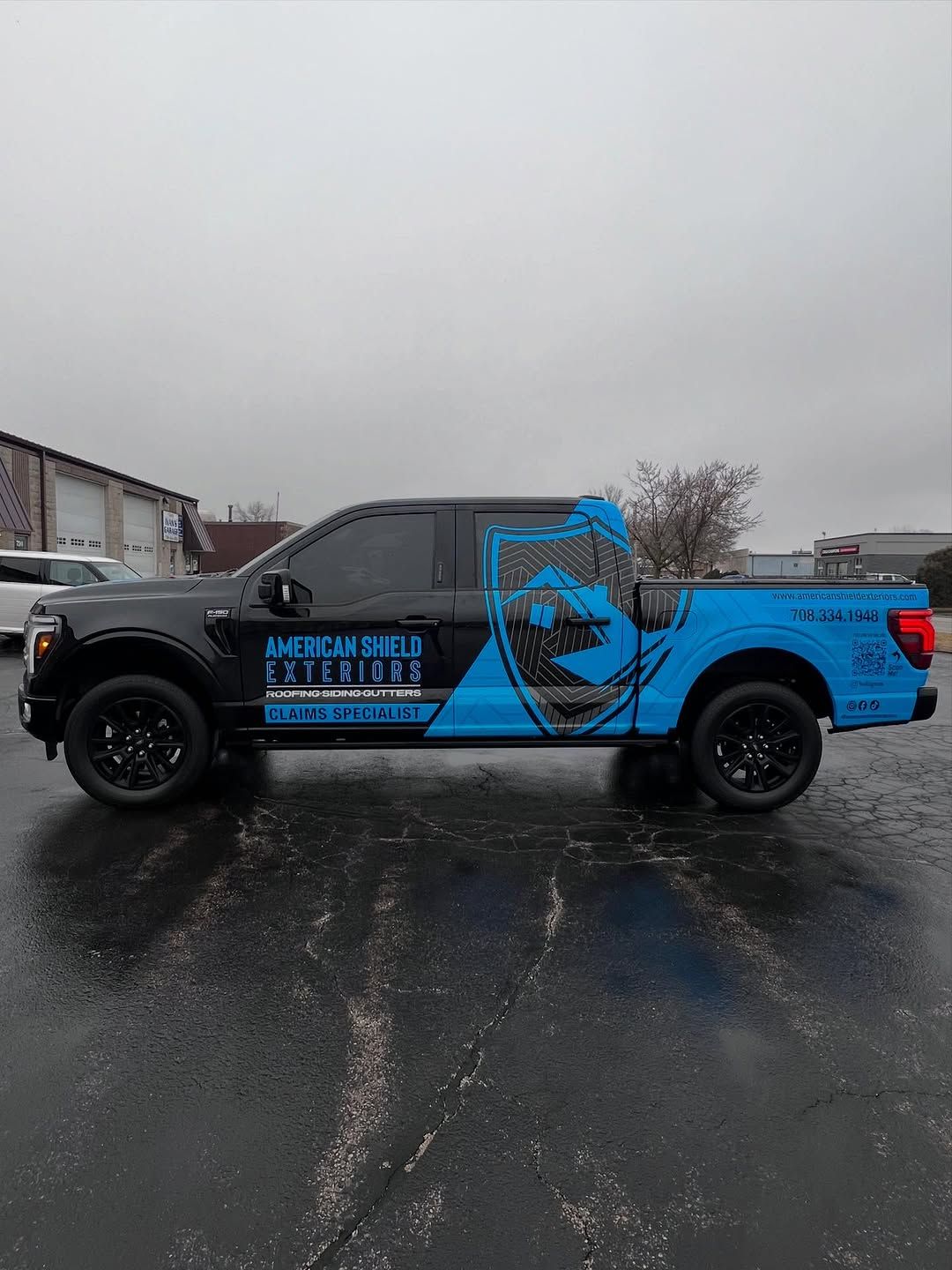 Black pickup truck with a blue and black partial wrap and American Shield Exteriors lettering on the door, in a wet lot.