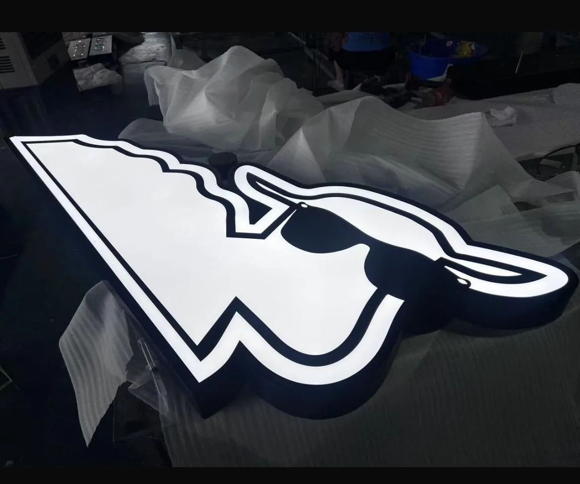 Illuminated white logo sign shaped as an arrow wearing sunglasses, lit on the shop floor before install.