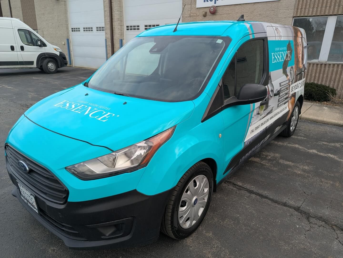 Teal Ford Transit Connect van in a full Essence wrap, parked in front of a row of garage doors.