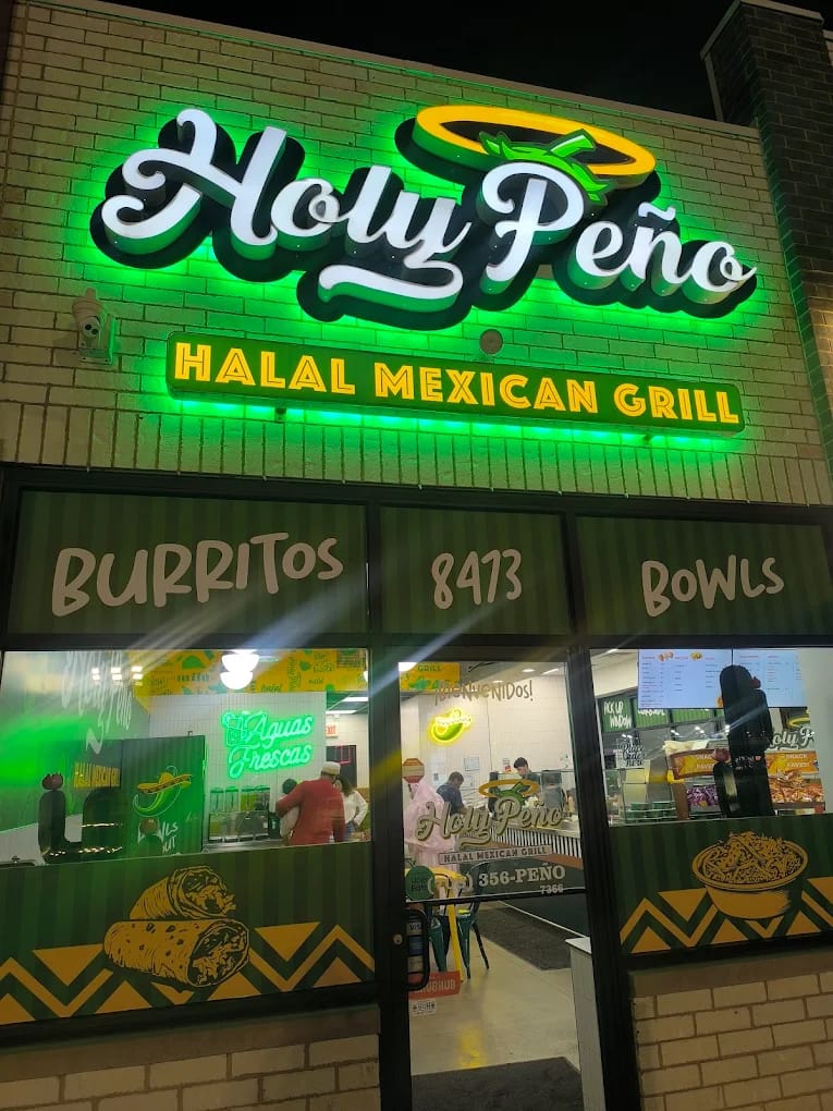 Holy Peño storefront at night: green illuminated script letters and a yellow HALAL MEXICAN GRILL sign above printed window graphics.