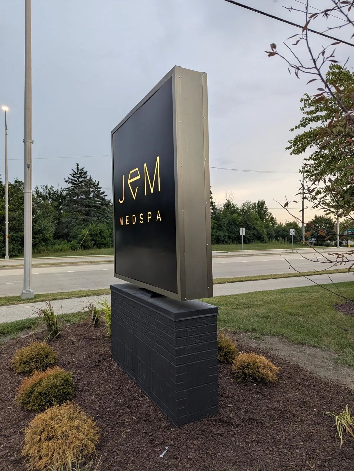 Brushed metal JEM Medspa monument sign on a dark base beside the road, with trees and a lawn behind it.
