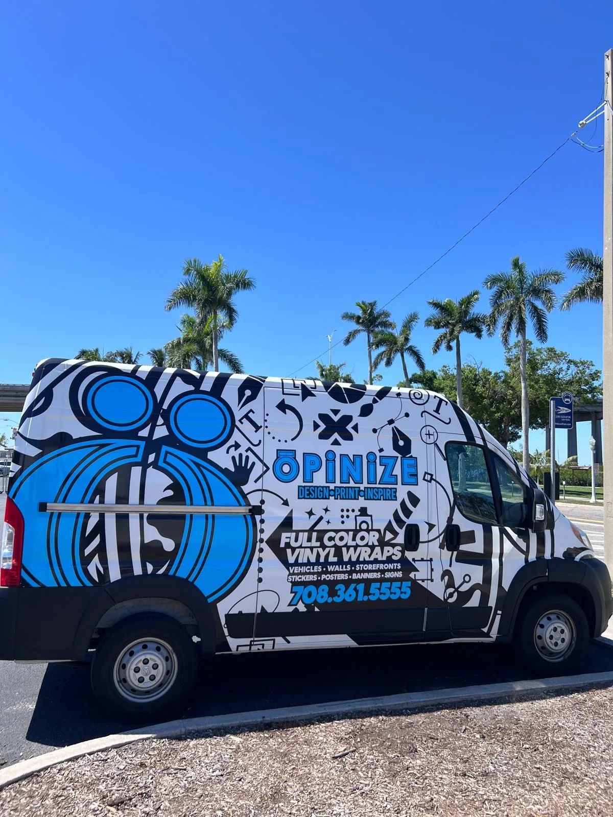 The shop's own cargo van in a blue Opinize wrap, FULL COLOR VINYL WRAPS and the phone number on the side, under palm trees.