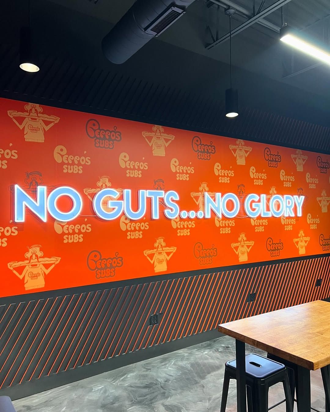 Orange printed wall inside a sandwich shop with a repeating Peppo's Subs mascot pattern and NO GUTS NO GLORY in blue neon-style lettering.