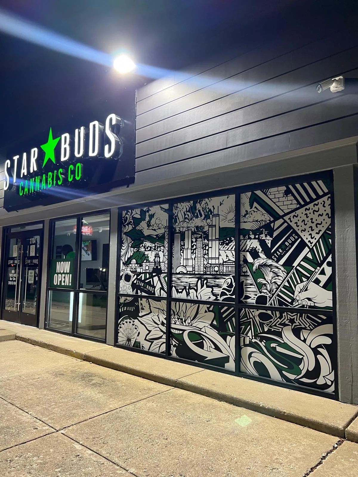 Star Buds storefront at night: the illuminated sign over the door and black and white printed graphics across the windows.