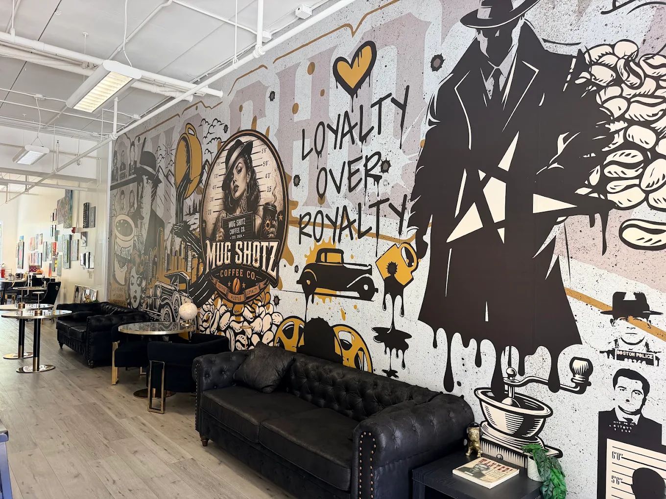 Illustrated gold and black mural inside Mug Shotz Coffee Co: a gangster in a trench coat, a mugshot portrait logo and the words LOYALTY OVER ROYALTY behind leather sofas.