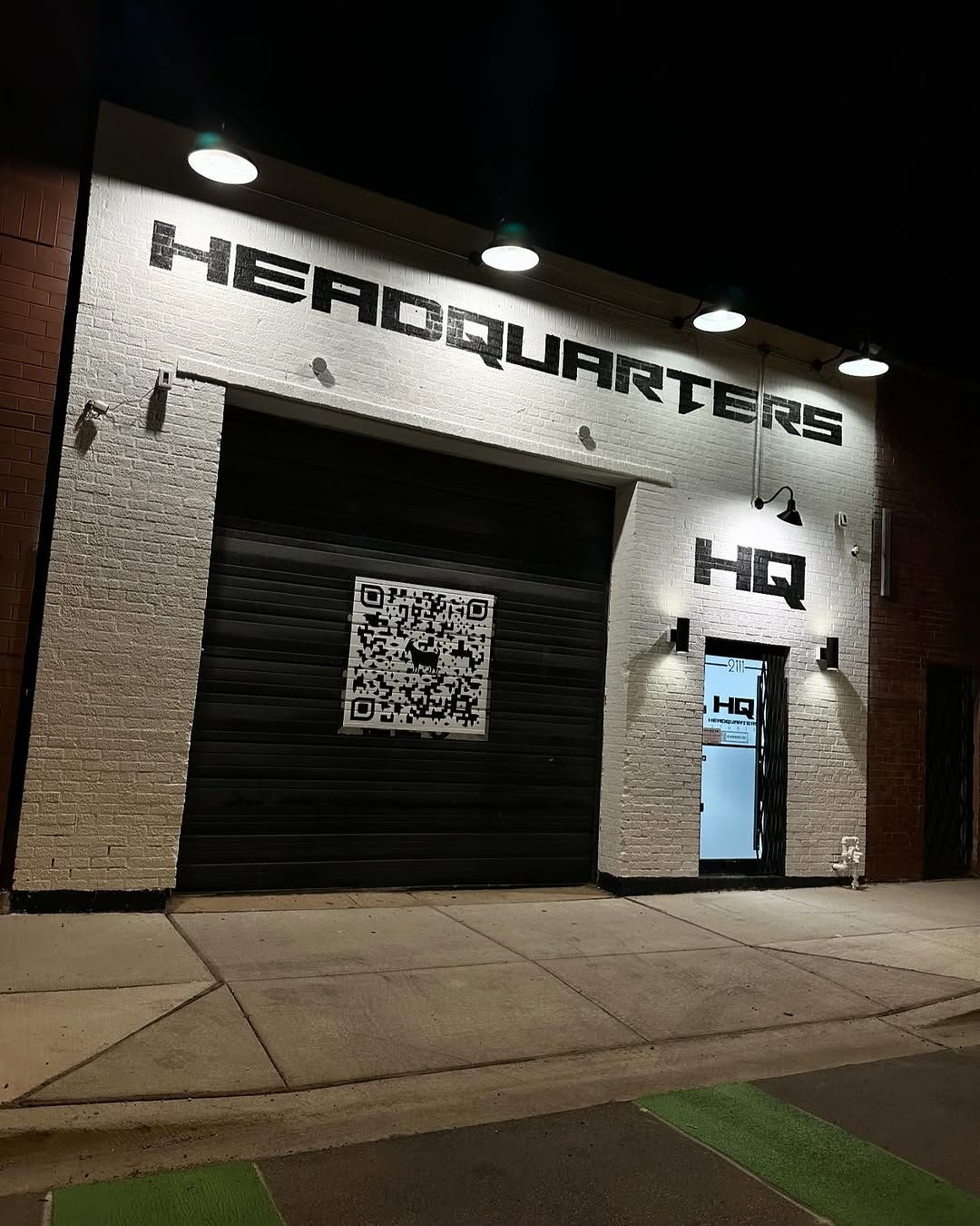 Big black HEADQUARTERS lettering across a white brick building front at night, lit by wall lights, with an HQ mark beside the door.