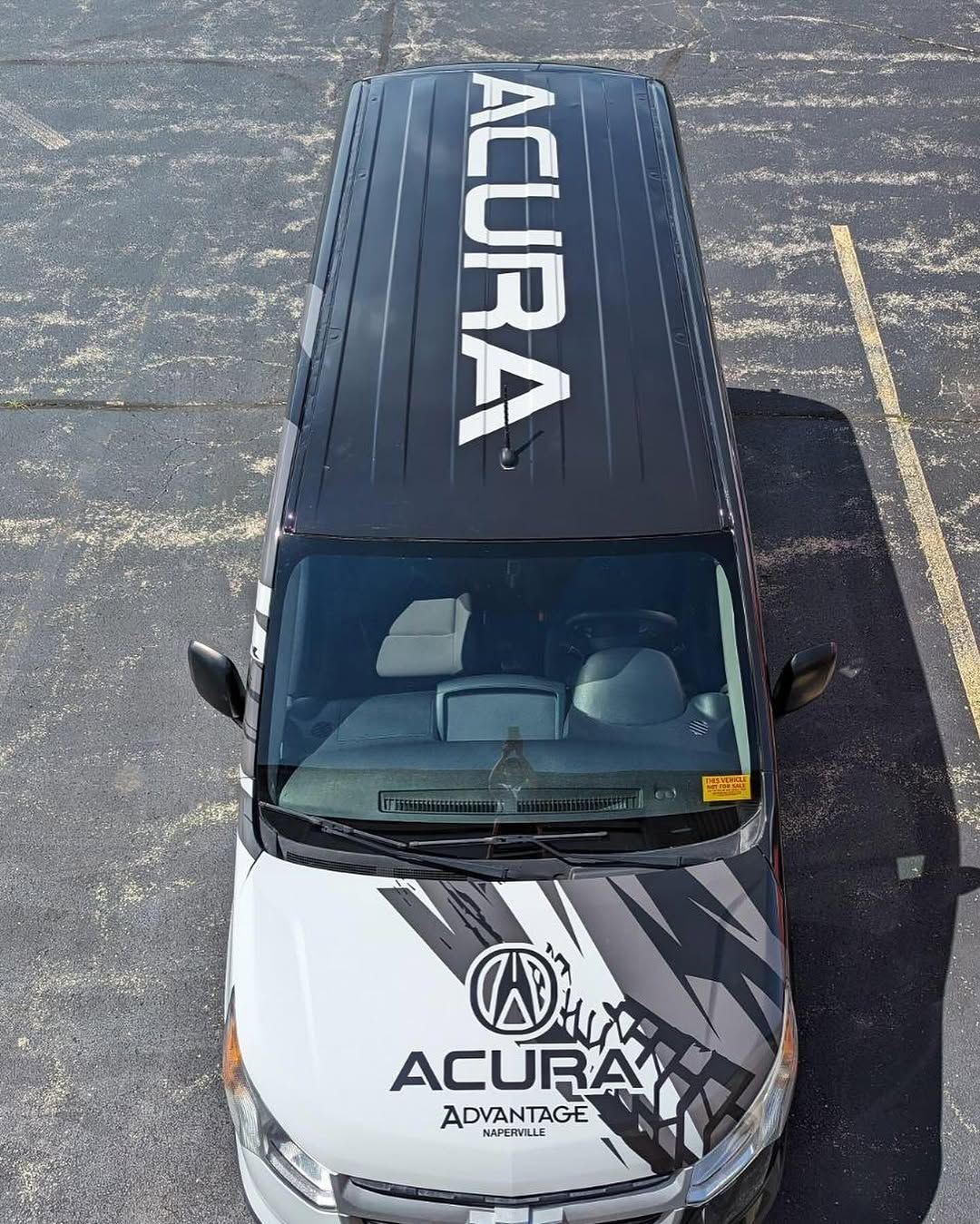 Black Acura SUV seen from above in a parking lot, with a large white ACURA graphic across the roof and a printed hood wrap.