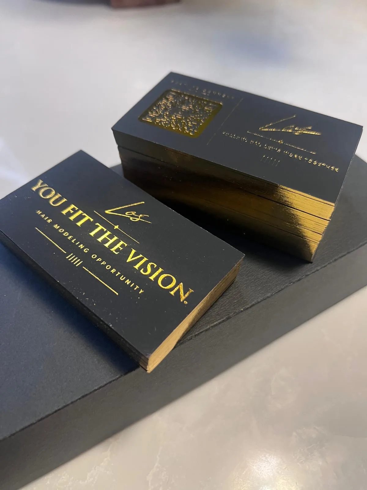 Two stacks of black business cards with gold foil lettering reading YOU FIT THE VISION, on a grey counter.