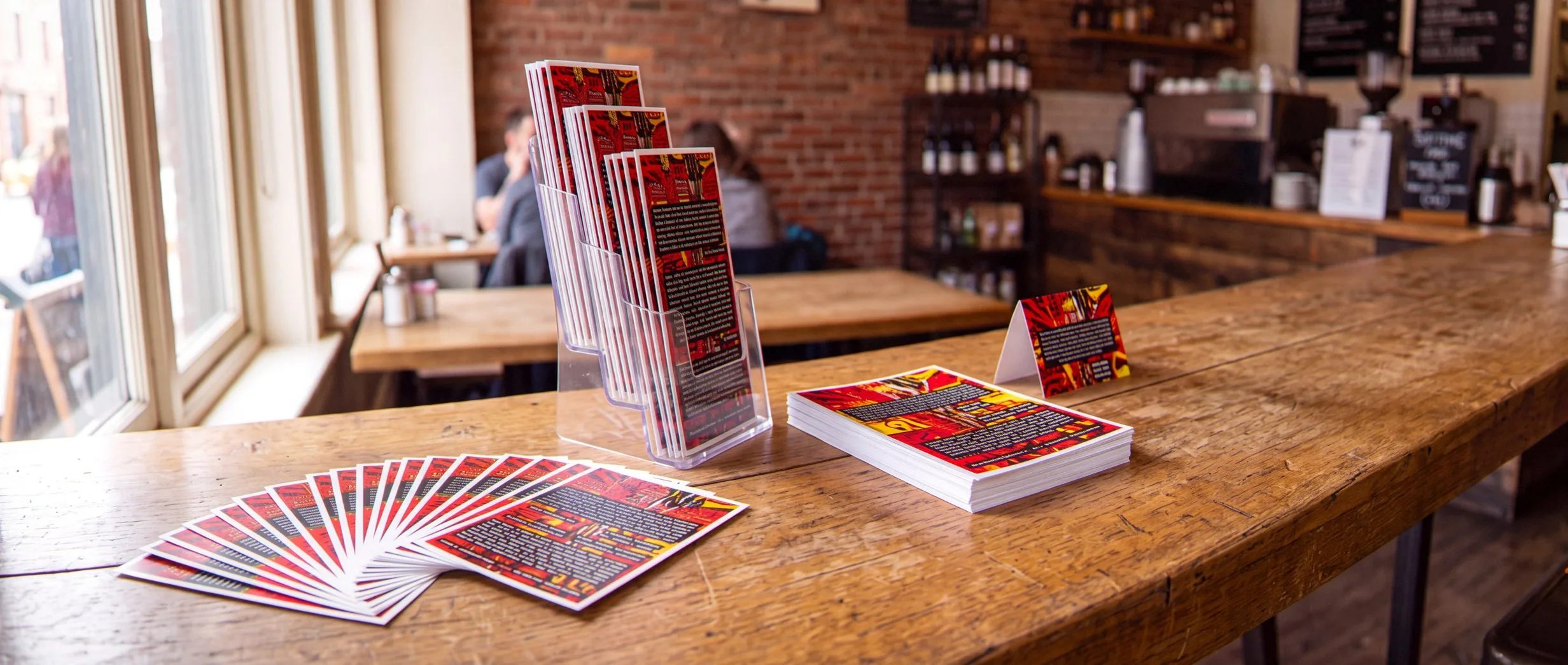 Fan of glossy postcards, a stack of flyers, folded brochures in a clear rack and a table-tent card along a café counter, all in red, black and yellow.